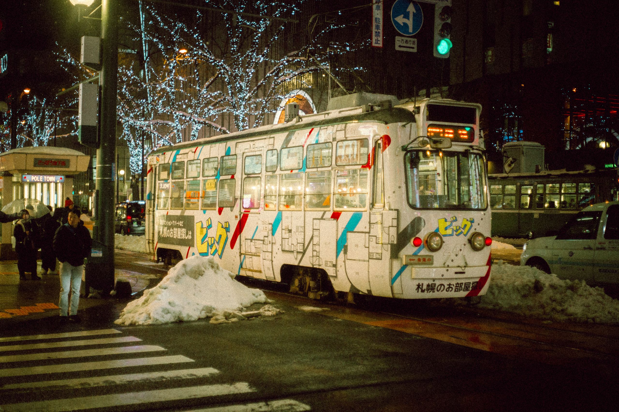 A vibrant tram travels through a snowy urban street, illuminated by festive lights at night.